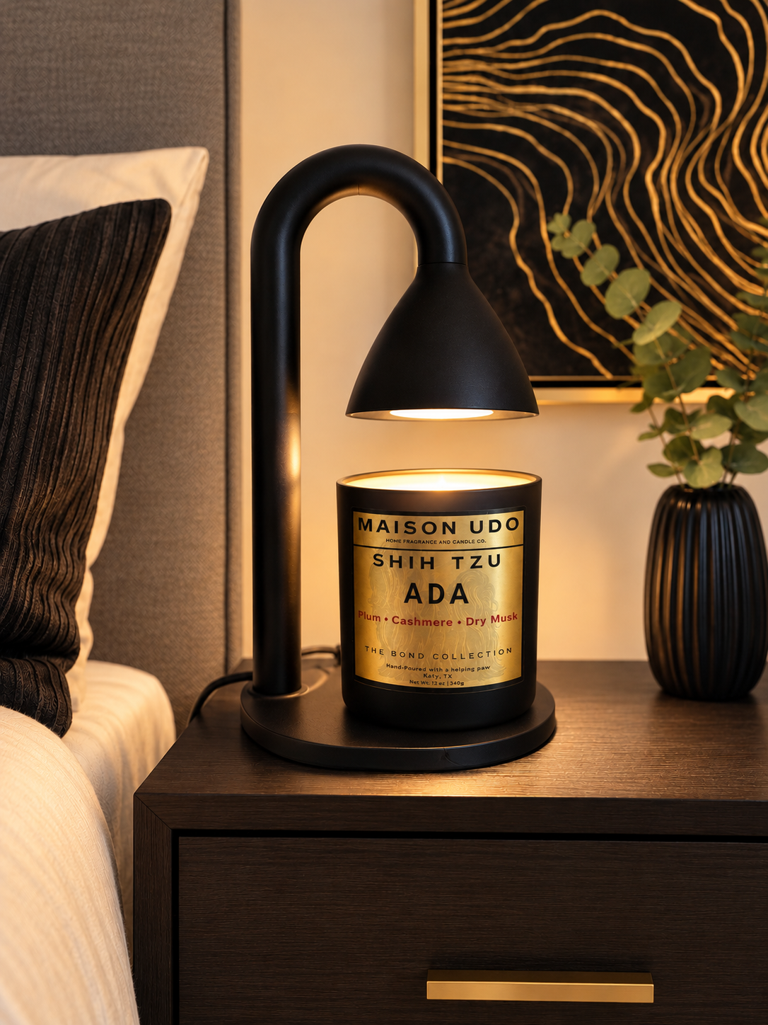 A black modern table lamp with an arched arm and dome shade sits on a dark wood nightstand, illuminating a Maison Udo candle. The candle is wrapped in gold and black paper with the text 'MAISON UDO SHIH TZU ADA'.